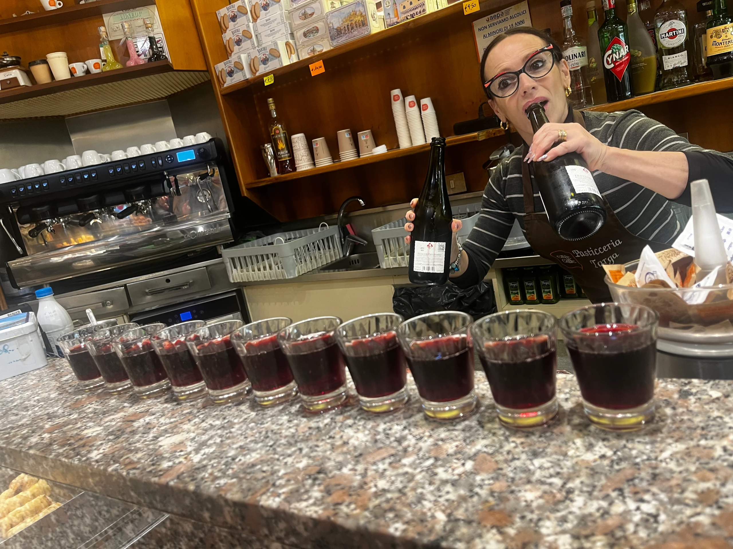A bartender at Pasticceria Targa in Venice pouring glasses of red wine and preparing pastries for a guided cicchetti tour group.